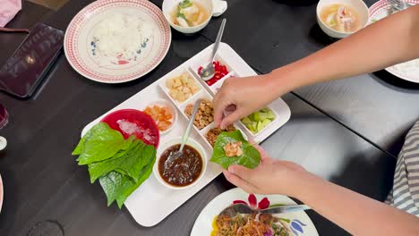 hands prepare traditional miang kham by adding assorted fillings to betel leaves on a white tray, under bright, even lighting, viewed from overhead
