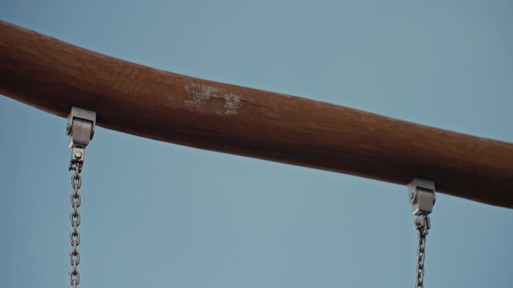 Wooden beam with metal hardware and chains sways gently against clear blue sky. camera slowly pans to reveal texture of weathered wood visible grain patterns small cracks or damage on its