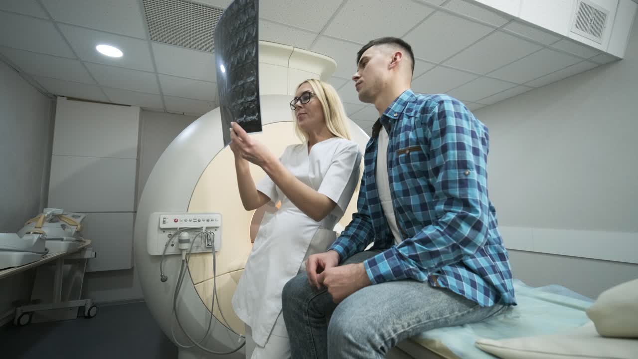 una mujer radióloga explica los resultados de la resonancia magnética para un paciente joven, mostrando la instantánea con imágenes, observando y analizando la resonanza magnética en una clínica moderna junto a una moderna máquina de escáner mri de tipo cerrado.