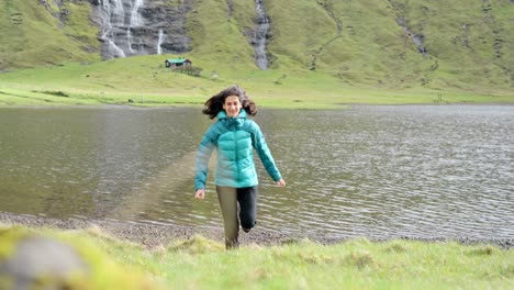 a woman in a blue jacket runs near a peaceful lake with grassy hills in faroe islands
