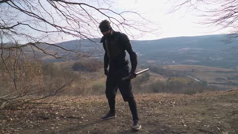a masked ninja warrior assumes a battle-ready stance on a hilltop, overlooking a vast valley. dressed in black tactical gear, he prepares for combat, embodying stealth and discipline
