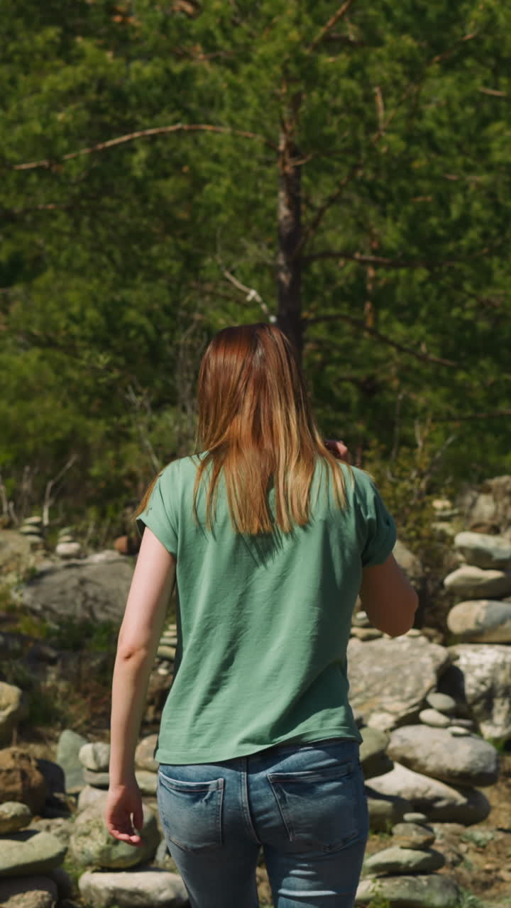 Woman walks between symbolic old stone piles near fir forest backside view slow motion. Place to meditate and rest at highland. Mystical traditions