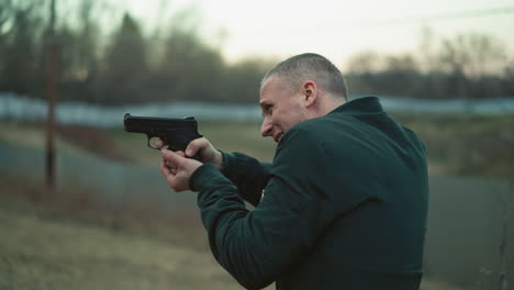 a close view of a man aiming a handgun with intense focus and determination, the outdoor setting includes a blurred background of a fence and a calm landscape