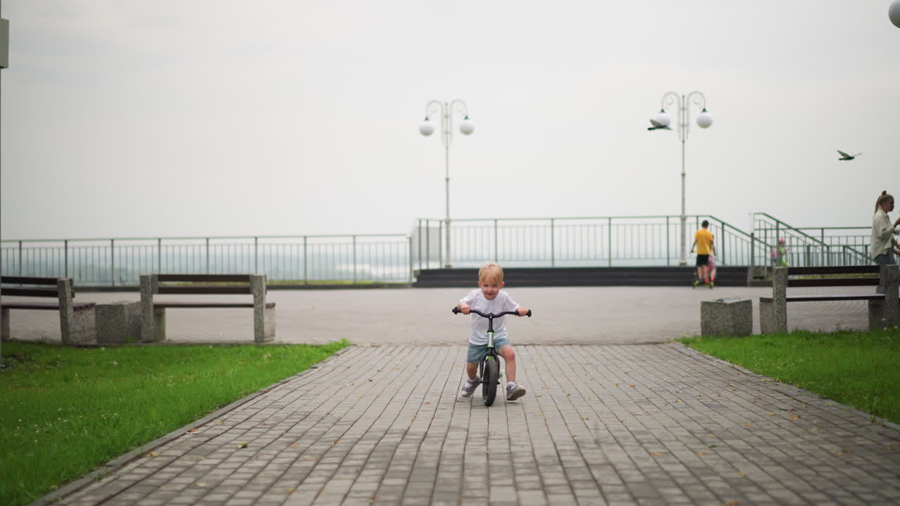 A happy little boy rides his bicycle down a paved path with a big smile, in the blurred background, other children can be seen walking