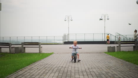 a happy little boy rides his bicycle down a paved path with a big smile, in the blurred background, other children can be seen walking