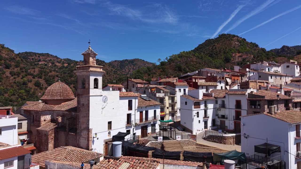 Villaggio tradizionale spagnolo di Chovar con edifici bianchi, tetti in terracotta e storica torre campanaria, immerso tra verdi colline boscose sotto un cielo azzurro e limpido