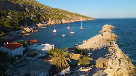 Yates-En-La-Bahía-Cerca-De-La-Ciudad-Costera-De-Croacia-Con-El-Telón-De-Fondo-De-Cielos-Azules-Y-Agua-Azul-Transparente,-árboles-Verdes-Y-Casas-Con-Techos-Rojos