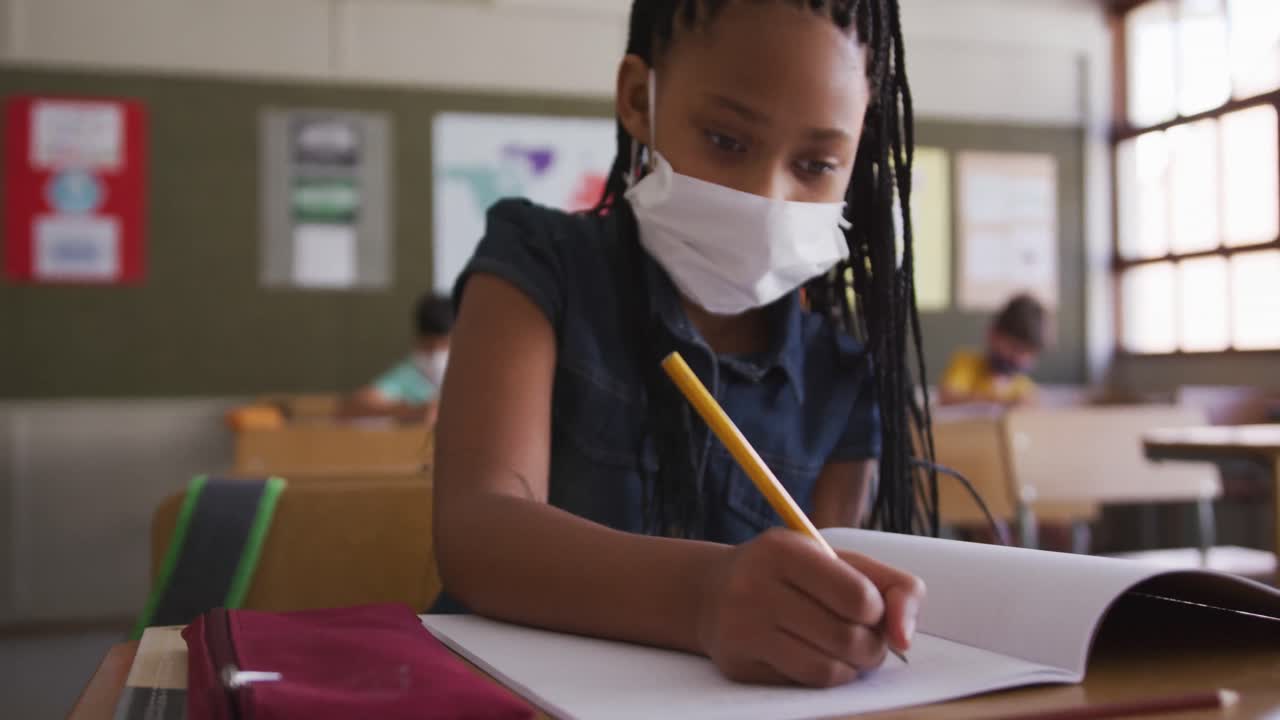 Girl wearing face mask writing while sitting on her desk at school