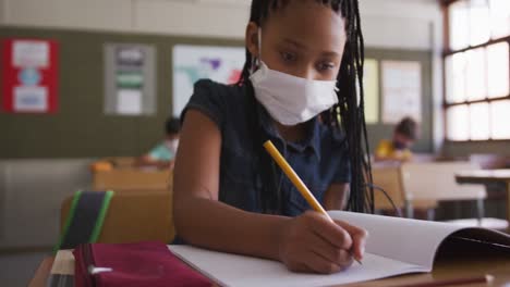 girl wearing face mask writing while sitting on her desk at school