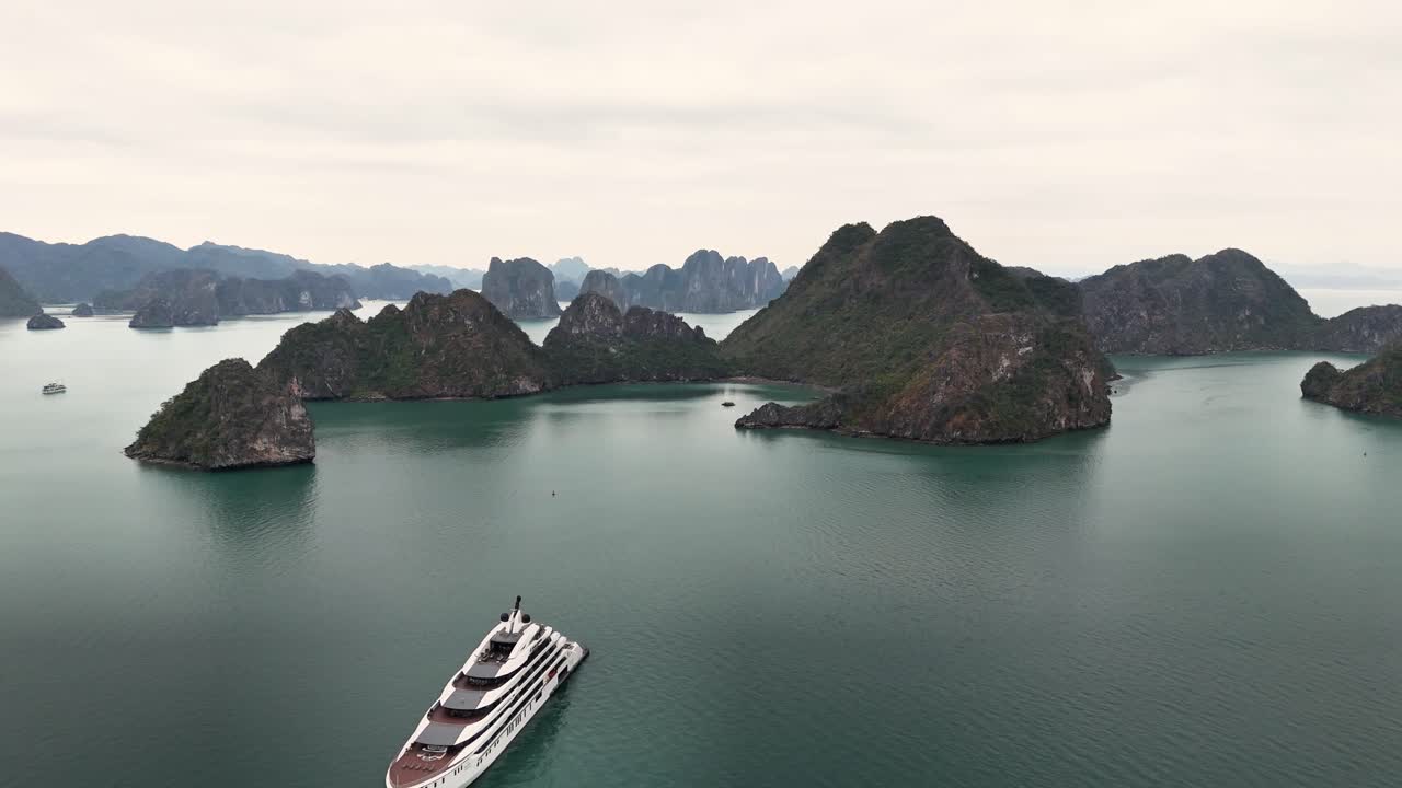 A luxury yacht floats in the calm turquoise waters of Ha Long Bay, Vietnam, encircled by towering limestone karst islands under a cloudy sky, creating a serene and picturesque seascape