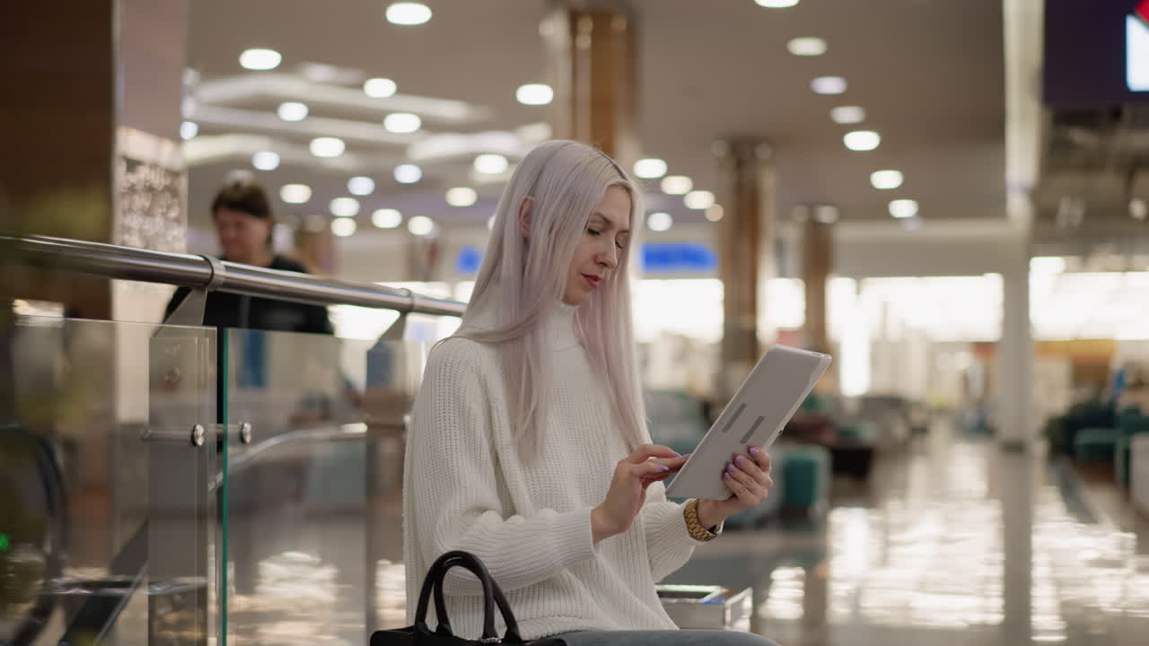 youthful lady seated on bench in spacious mall corridor wearing white sweater and jeans, engrossed in tapping tablet with focused expression, black handbag beside, modern interior lighting