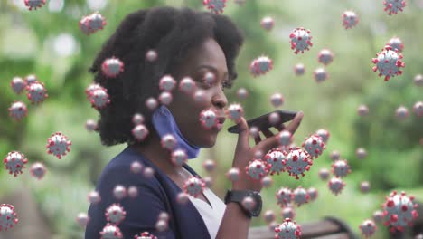 Animation-of-coronavirus-cells-over-african-american-woman-with-face-mask-talking-on-phone-in-park