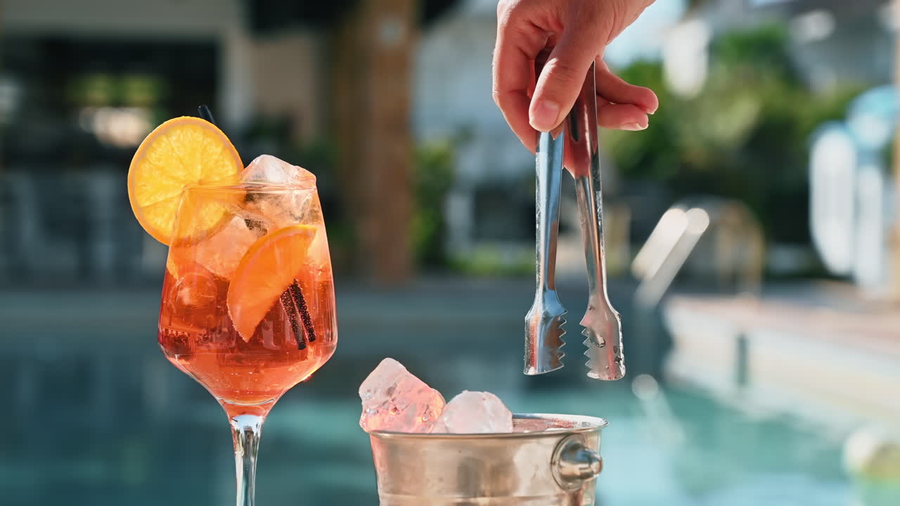 Cocktail e secchiello con ghiaccio, fetta d'arancia, uomo che aggiunge ghiaccio usando le pinze, piscina sullo sfondo, Grecia. Rallentatore