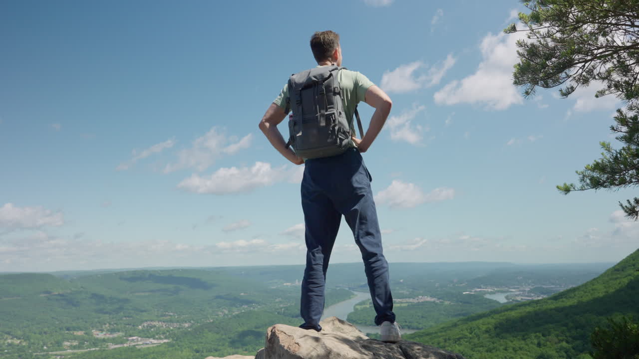 Young man successfully reaches top of a mountain after long climb. Stands confidently and proudly on a rock overlooking the amazing view and natural landscape. Green plains and forest trees and river