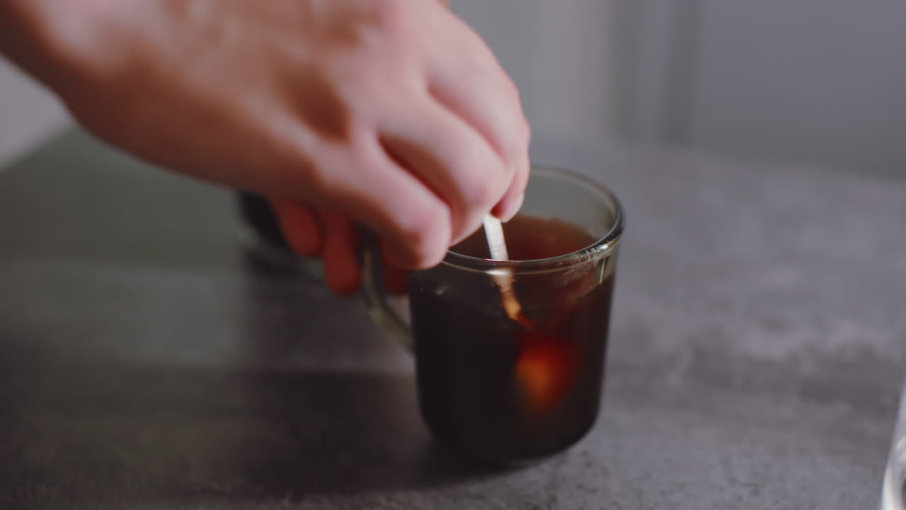 Primo piano di una mano che mescola del caffè appena fatto in una tazza di vetro trasparente, agitando il liquido scuro con un cucchiaino, creando un movimento circolare mentre prepara una bevanda calda per rinfrescarsi al mattino sul tavolo della cucina