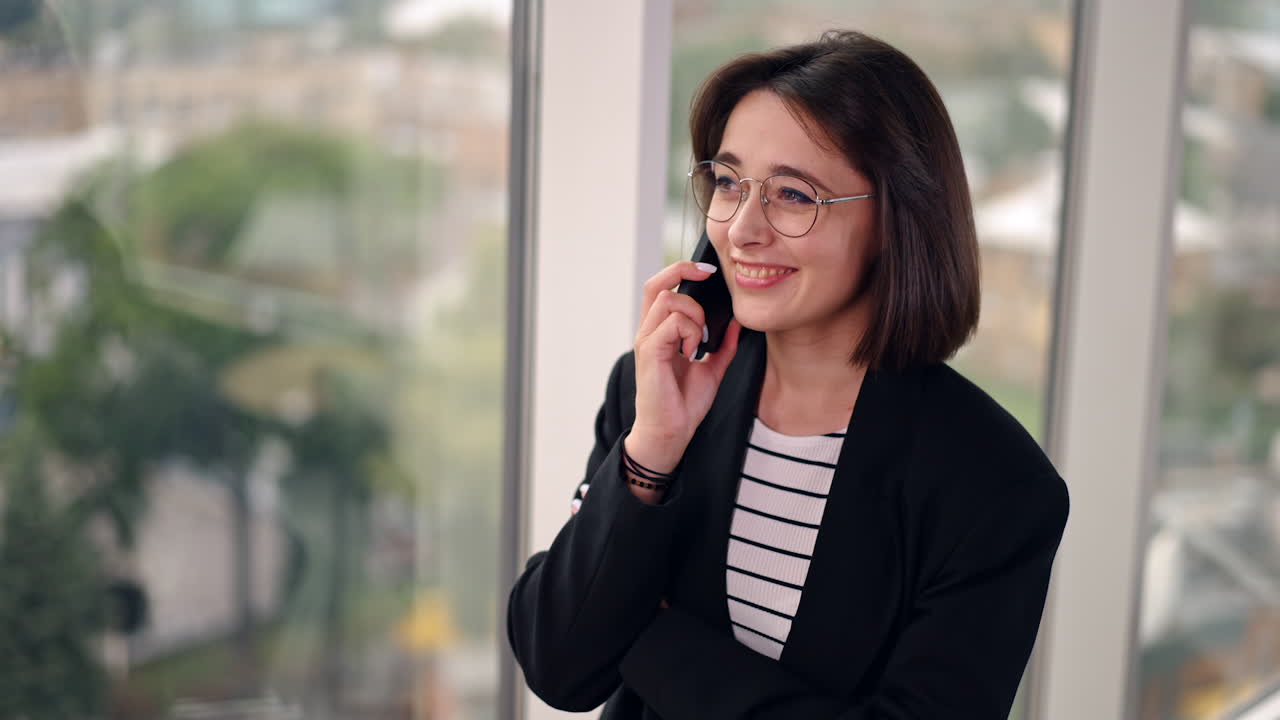 Attractive Caucasian lady wearing glasses and black jacket stands at the window. Woman speaks on the phone smiling.