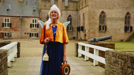 a happy woman dressed as a fairy talking towards camera while standing in front of a medieval castle