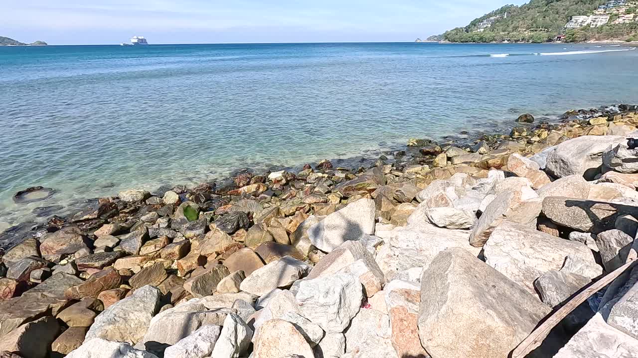 A tranquil view of Kalim Beach's rocky shoreline under clear skies, showcasing calm waters and distant hills in Phuket, Thailand