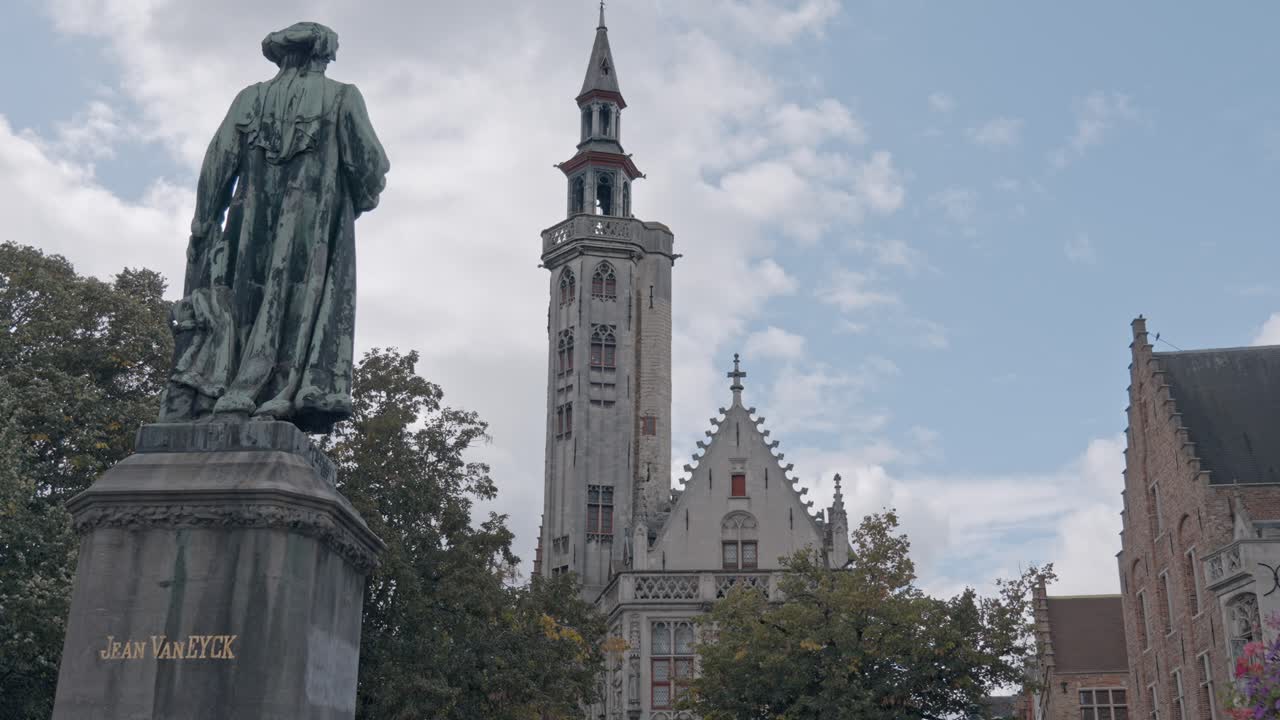A bronze statue of the Flemish master painter Jan van Eyck stands prominently in Jan van Eyckplein (Jan van Eyck Square) in Bruges, Belgium. In the background, tower of the Poortersloge