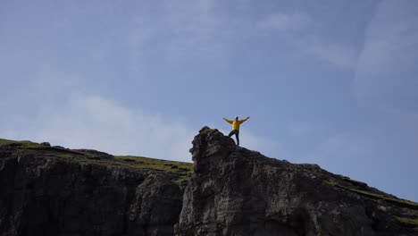 Man-in-yellow-raincoat-stands-victorious-on-top-of-Traelanipa-sheer-cliff-against-blue-sky,-Faroe-Islands