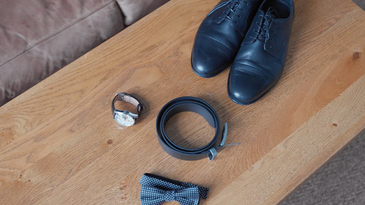 Stylishly arranged men's accessories including a belt, watch, and bow tie on a wooden table. The scene is accented by leather shoes, providing a classic, elegant vibe