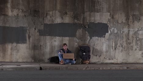 Wide-Shot-of-a-Homeless-Man-Sitting-on-the-Sidewalk-Holding-a-Cardboard-Sign