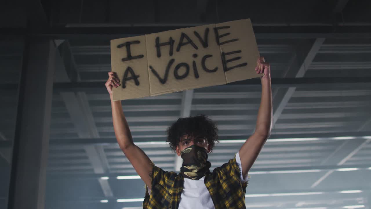 hombre de raza mixta con máscara facial sosteniendo una pancarta de protesta en un garaje vacío