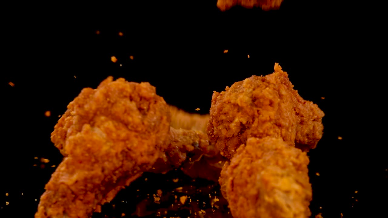 Golden fried chicken pieces tumble and land against a black background, illuminated by dramatic lighting, with crumbs and seasoning scattering in slow motion