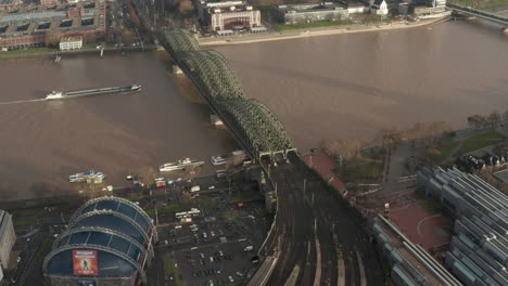 Aerial-footage-of-cargo-boat-passing-on-wide-Rhine-river-near-railway-bridge.-Heavy-traffic-on-waterfront-road.-Cologne,-Germany