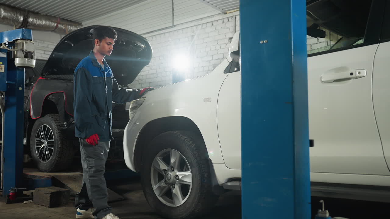mecánico en uniforme azul revisa el neumático del coche golpeándolo con la pierna en el taller de automóviles, realizando la inspección y mantenimiento del vehículo en un centro de servicio mecánico