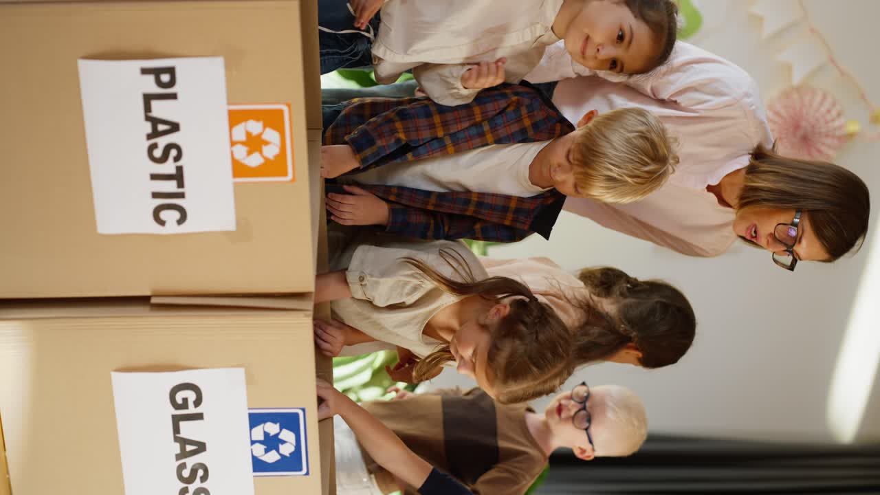 Vertical video of a woman teacher in a pink shirt with glasses with a bob hairstyle teaching them together with preschool children how to properly sort glass, plastic and paper in a special box in a club for preschool children preparing for school