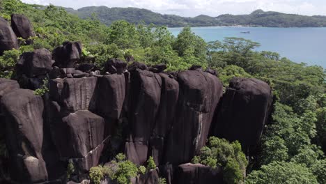 Drone-reveal-footage-of-huge-granite-stones-on-mountain-top-near-the-beach,-surrounded-by-trees,-Mahe-Seychelles-30-fps