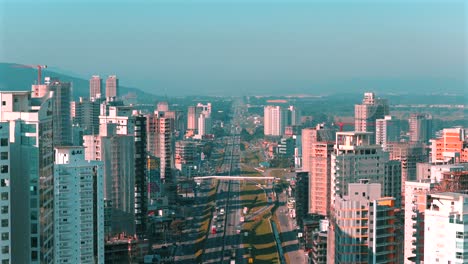 descending aerial view of tall residential buildings flanking the main highway running through itapema city, brazil, with distant hills and blue sky defining the landscape of santa catarina