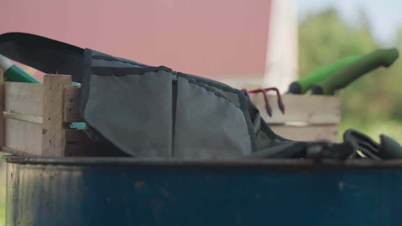 Close up male worker removing apron belt from waist, dropping strap onto wooden crate beside blue drum with gloves and hand tools, backyard house fence in background