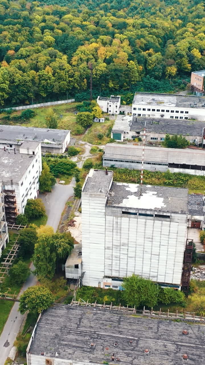 Empty factory constructions. Flight over abandoned buildings with old roofs. Ruined industrial territory near the forest.. Vertical video