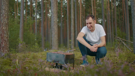 un hombre adulto alegre está sentado cerca de una parrilla con carne y salchichas en el bosque sonriendo y disfrutando del descanso en la naturaleza