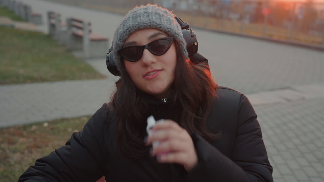 Giovane donna con le cuffie su una panchina che svapa al tramonto, sorride e soffia fumo verso la telecamera, indossa occhiali da sole e un berretto di lana, con un ritmo rilassato e un sottile ballo a tempo di musica, in un'atmosfera cinematografica di luce solare.