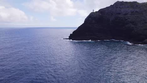 Drone-footage-approaching-Makapu'u-Point-Lighthouse-on-top-of-a-steep-cliff-overlooking-the-blue-Pacific-Ocean-water-with-white-capped-waves-crashing-onto-the-rocky-shore-below-Oahu-Hawaii