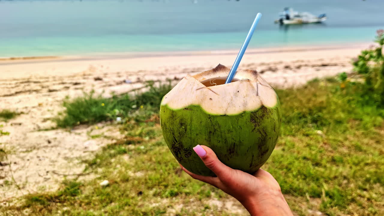 Hand holds coconut drink on pink beach in Lombok, relaxing tropical scene