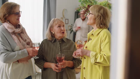 senior female friends holding wine and chatting at home party