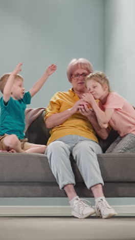 little children, brother and sister, sit with grandmother on couch, who holds piggy bank in hands, girl tries to take piggy bank, boy jumps sitting on sofa in room, next to lamp and flower