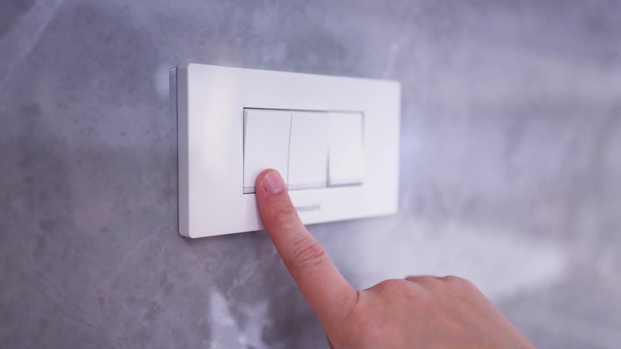 A hand presses a white wall switch, turning on warm indoor lighting against marble