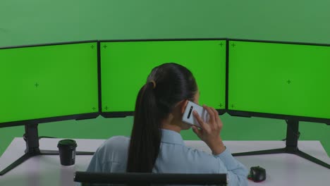 녹색 스크린 스튜디오에서 여러 컴퓨터 모니터를 모방하여 작업하는 동안 휴대 전화로 대화하는 아시아 여성의 뒷면
