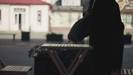 remote worker prepares to sit at an outdoor cafe, placing her bag on a table, the blurred background highlights urban elements, including buildings, poles, and flowers