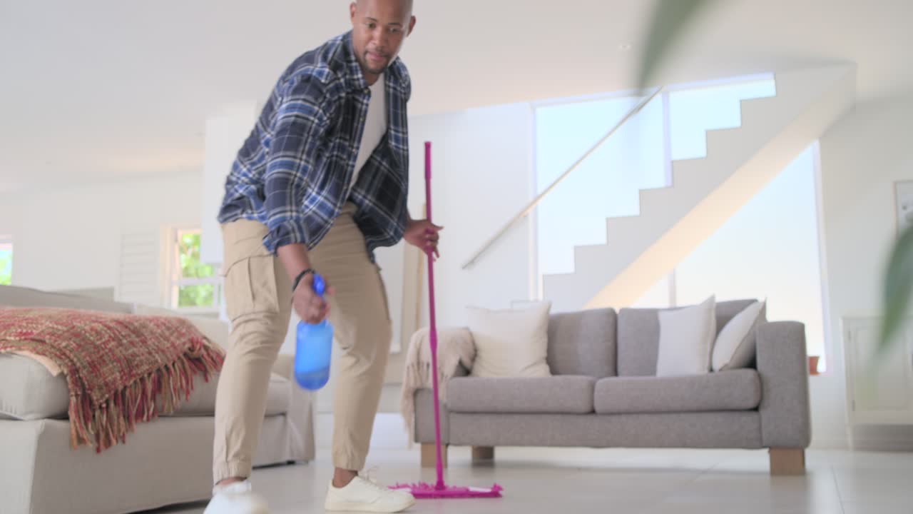 Afroamerikansk mann kommer inn, tar frem mopp, sprayer gulvet og vasker det hjemme, kledd i rutete skjorte.