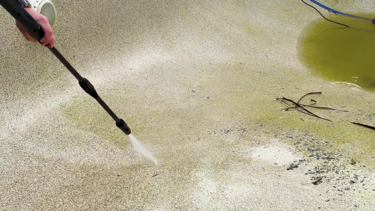 En højtryksrenser fjerner grønne alger og snavs fra en betonpooloverflade i slow-motion.