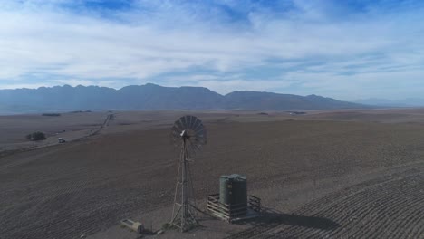 Die-Drohne-Fliegt-Im-Trockenen-Sommer-über-Trockenes-Ackerland-Und-Dreht-Die-Windpumpe