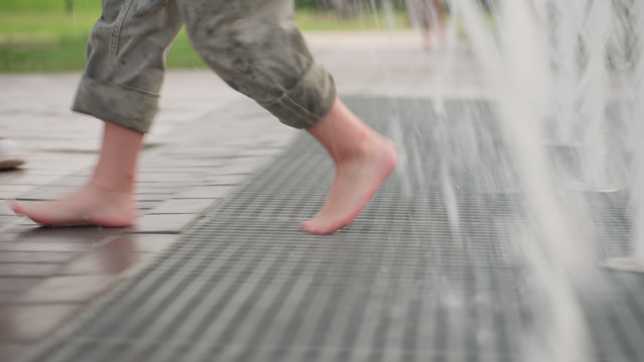 boy running barefoot across splash fountain, leg view showing motion and water droplets scattering on mesh surface, summer park playful energy, wet grid texture underfoot, casual childhood fun