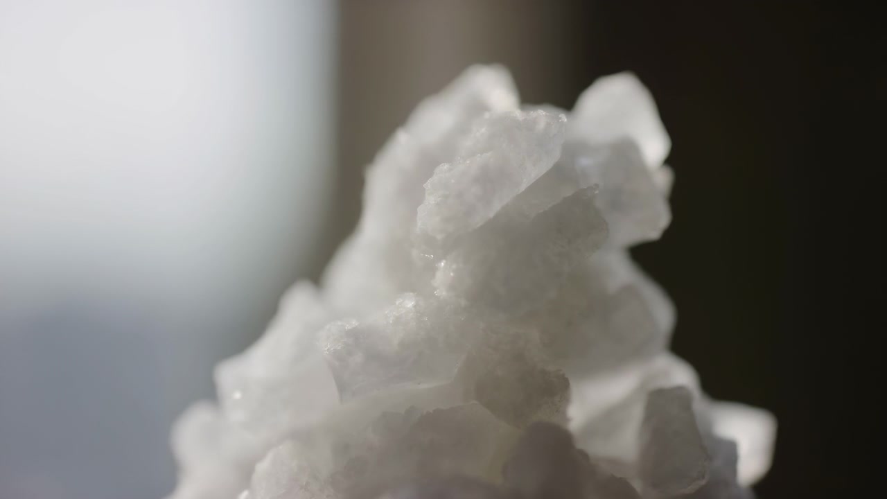 Un primo piano macro in movimento mostra una cima di un cumulo cristallino bianco in primo piano a destra che diventa sottilmente più prominente mentre le facce superiori acquisiscono definizione dei bordi e riflessi speculari più luminosi, indicando una leggera messa a fuoco