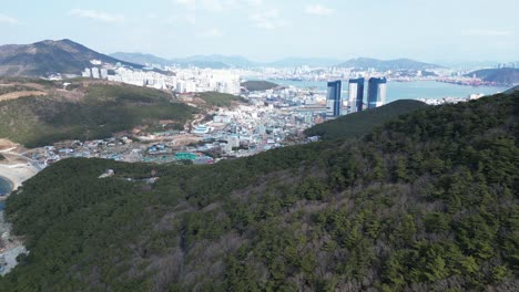 drone aerial view in south korea countryside flying over a green and rocky mountain next to dark blue sea of busan and city in the background sunny day